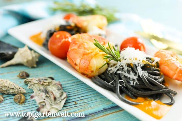 Große Garnelen, Cocktail-Tomaten und schwarze Spaghetti sowie Hummersauce befinden sich auf einem weißen länglichen Teller. Der Untergrund besteht aus blauen Brettern. Darauf sind Muscheln verteilt.