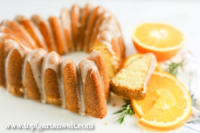 Orangenkuchen in Form von einem Herz. Die Mehlspeise ist angeschnitten. Ein Stück liegt daneben. Im Hintergrund sind Orangenhälften zu sehen.