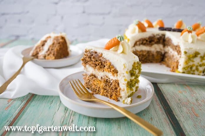 Eine Rüblitorte mit Frischkäse-Frosting steht angeschnitten auf einem weißen Teller. Sie ist mit den typischen Marzipanmöhren verziert. Daneben liegen goldene Gabeln.