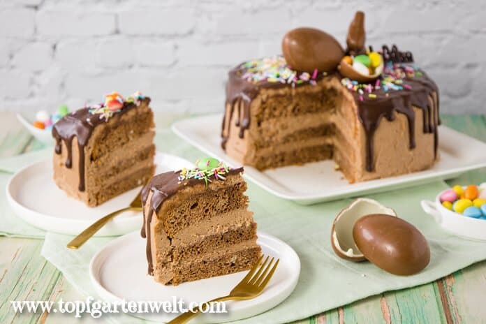 Die Ostertorte verziert mit Schoko-Drip, Überraschungsei und einem Schokoosterhasen, steht angeschnitten auf einem Teller. Zwei Stücke sind daneben platziert. Man sieht, dass sie aus drei Böden mit Creme dazwischen besteht.