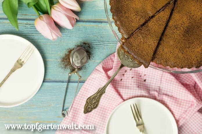 Eine Schokotarte bestreut mit Kakao auf einer Etagere wurde von oben fotografiert. Im Hintergrund liegen rosarote Tulpen. Daneben stehen zwei Teller mit goldenen Gabeln.