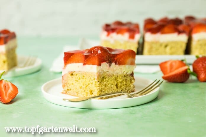 Ein Stück Erdbeerschnitte auf einem weißen Teller. Daneben liegt eine goldene Gabel. Im Hintergrund ist noch mehr vom Kuchen zu sehen.