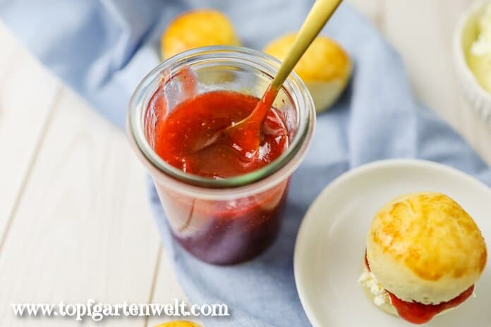 Ein Glas Erdbeermarmelade steht bereit. Daneben sind schon fertige Scones mit Clotted Cream zu sehen.