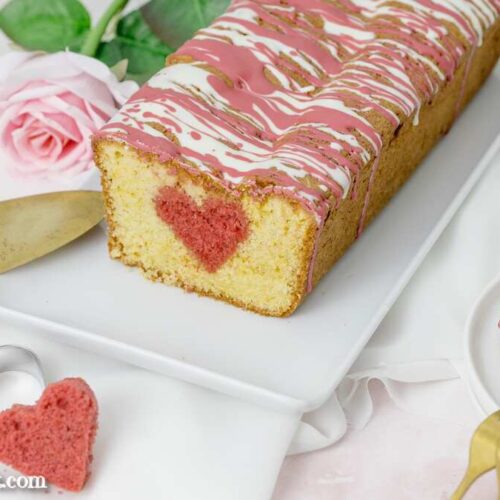 Ein einfacher Kuchen mit einem Herz in der Mitte auf einem weißen Teller. Daneben liegt eine goldene Kuchenschaufel. Im Hintergrund ist eine Rose zu sehen.