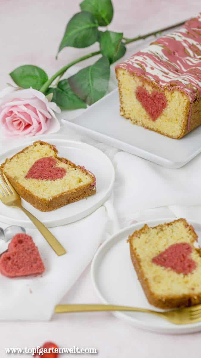 Ein Kuchen mit einem Herz in der Mitte ist im Hintergrund zu sehen. Davor sind zwei Teller auf denen jeweils ein Stück liegt. Daneben sind goldene Gabeln.