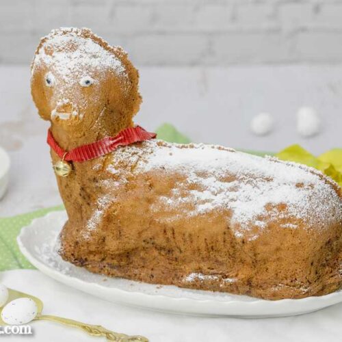 Ein Osterlamm aus Sandmasse steht auf einem weißen, ovalen Teller. Im Hintergrund sind gelbe Tulpen zu sehen.