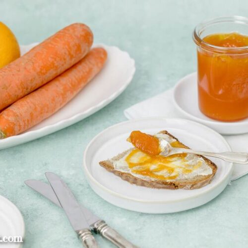 Ein Glas mit Ostermarmelade steht im Hintergrund. Davor liegt eine Scheibe Brot bestrichen mit Butter und dem Fruchtaufstrich. Als Steudeko sind kleine Eier und die Zutaten Möhren, Äpfel und Orangensaft zu sehen.