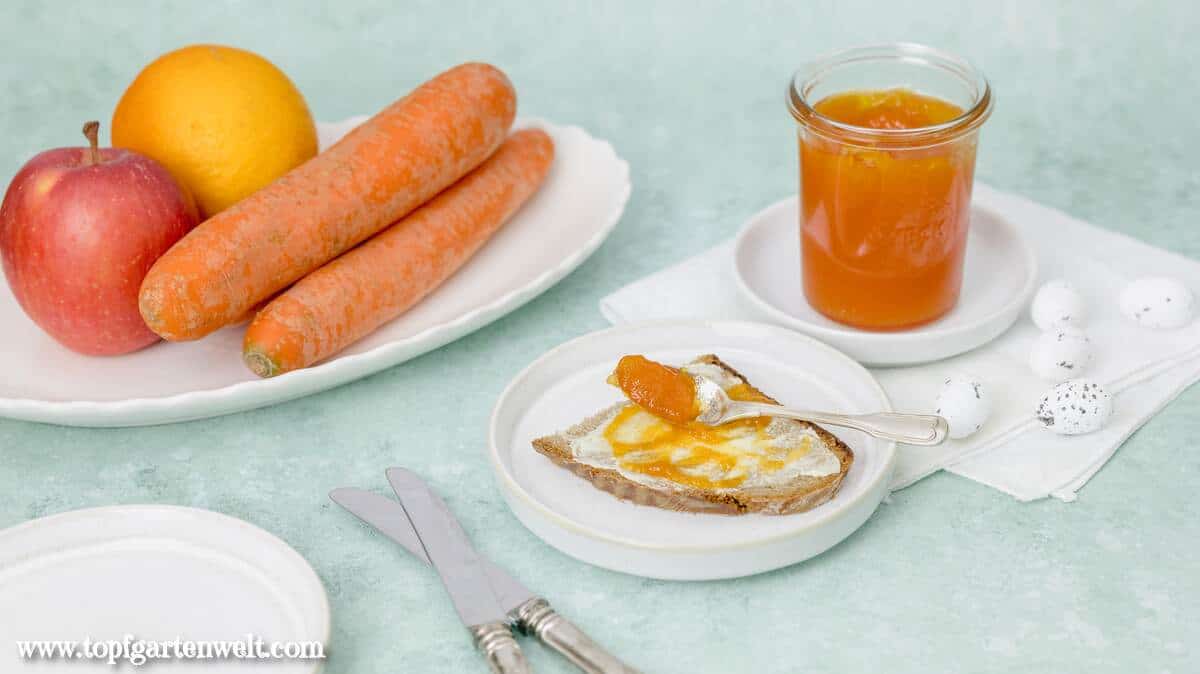 Ein Glas mit Ostermarmelade steht im Hintergrund. Davor liegt eine Scheibe Brot bestrichen mit Butter und dem Fruchtaufstrich. Als Steudeko sind kleine Eier und die Zutaten Möhren, Äpfel und Orangensaft zu sehen.