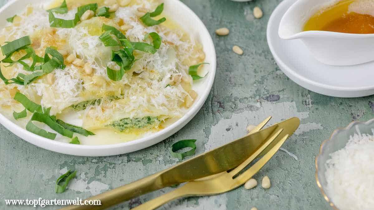 Ein Teller voll mit Bärlauch Ravioli garniert mit brauner Butter, Parmesan und Pinienkernen. Im Vordergrund liegt goldenes Essbesteck.