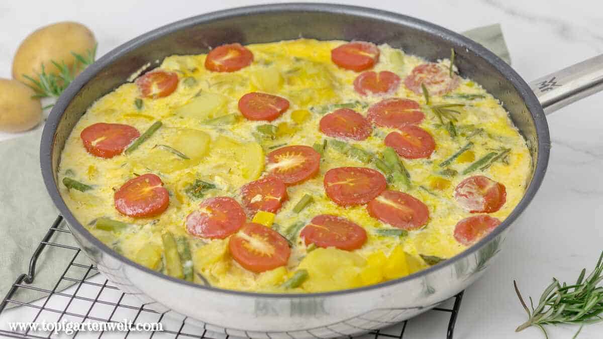 Frittata mit grünem Spargel und Kartoffeln in einer Pfanne, die auf einem schwarzen Abkühlgitter steht. Davor ist ein wenig frischer Rosmarin zu sehen.