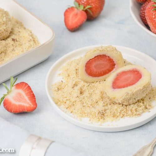 Zwei Hälften eines Erdbeerknödels sind auf Mandel-Butterbrösel angerichtet. Im Hintergrund sind weitere Früchte und Knödel zu sehen.