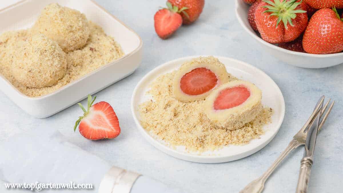 Zwei Hälften eines Erdbeerknödels sind auf Mandel-Butterbrösel angerichtet. Im Hintergrund sind weitere Früchte und Knödel zu sehen.