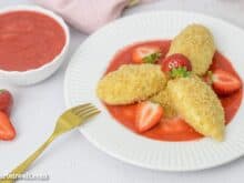 Drei Topfennockerl mit Erdbeersauce und Butterbrösel sind auf einem Dessertteller angerichtet. Im Hintergrund ist eine Schüssel mit Fruchtpüree zu sehen.