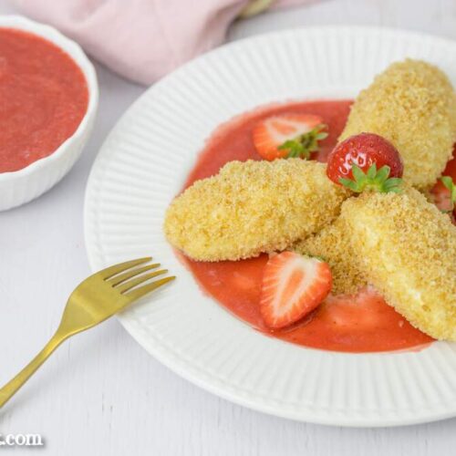 Drei Topfennockerl mit Erdbeersauce und Butterbrösel sind auf einem Dessertteller angerichtet. Im Hintergrund ist eine Schüssel mit Fruchtpüree zu sehen.