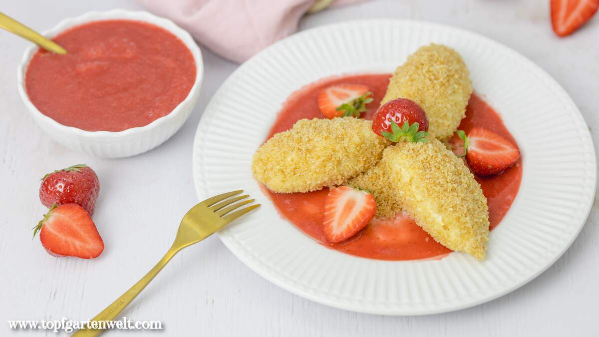 Drei Topfennockerl mit Erdbeersauce und Butterbrösel sind auf einem Dessertteller angerichtet. Im Hintergrund ist eine Schüssel mit Fruchtpüree zu sehen.