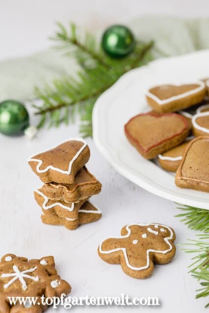 Vier Lebkuchenkekse sind hier über einander gestapelt. Sie sind mit weißem Zuckerguss verziert. Im Hintergrund ist ein Teller voll gefüllt mit Plätzchen zu sehen.