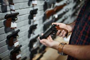 A person holds a pistol in a gun store with dozens of firearms on the wall before them.