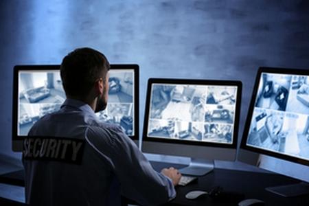 A male security guard sits in front of three computer monitors streaming live footage from inside a building.