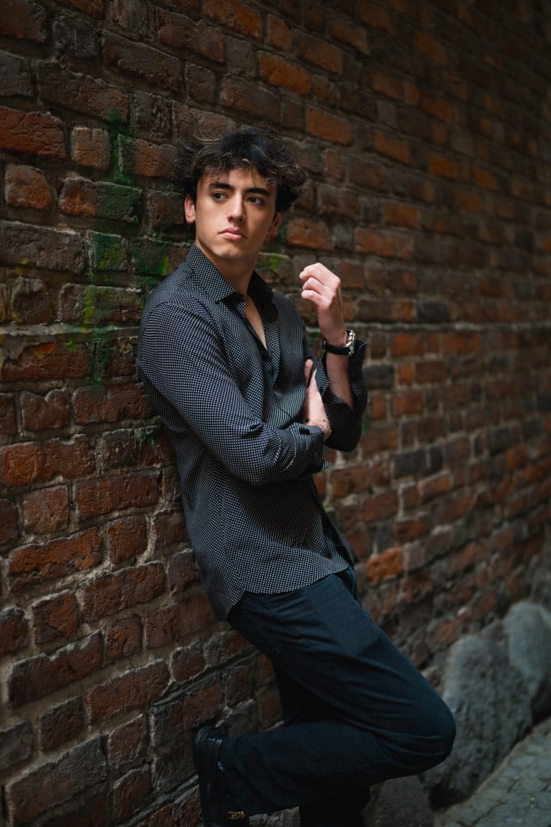A man in a black polka-dot shirt and dark trousers, leaning against an old brick wall. Tinder portrait.