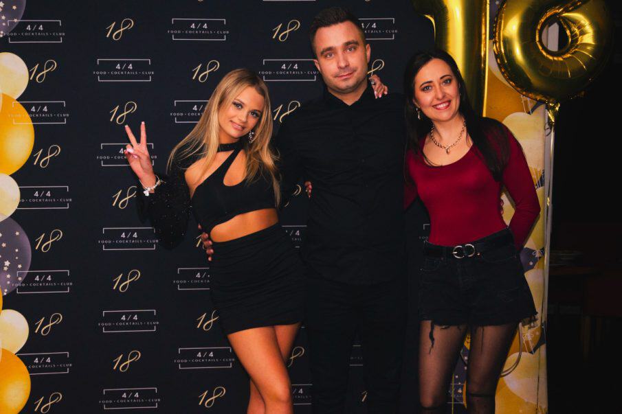 A formal group portrait at an 18th birthday party, featuring the birthday girl in black with guests against a custom "18" logo wall with balloons.