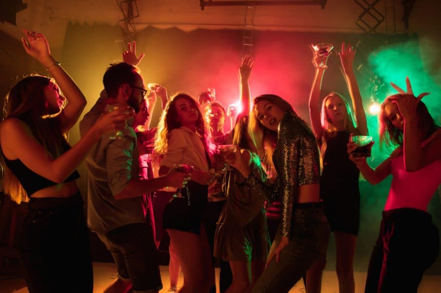 A group of young friends celebrating an 18th birthday, dancing with colorful drinks under vibrant party lights and atmospheric smoke.