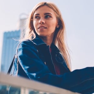 A satisfied young client with blonde hair in a denim jacket, standing against modern warsaw architecture. She recommends professional event photography and dynamic reportage services in warsaw.