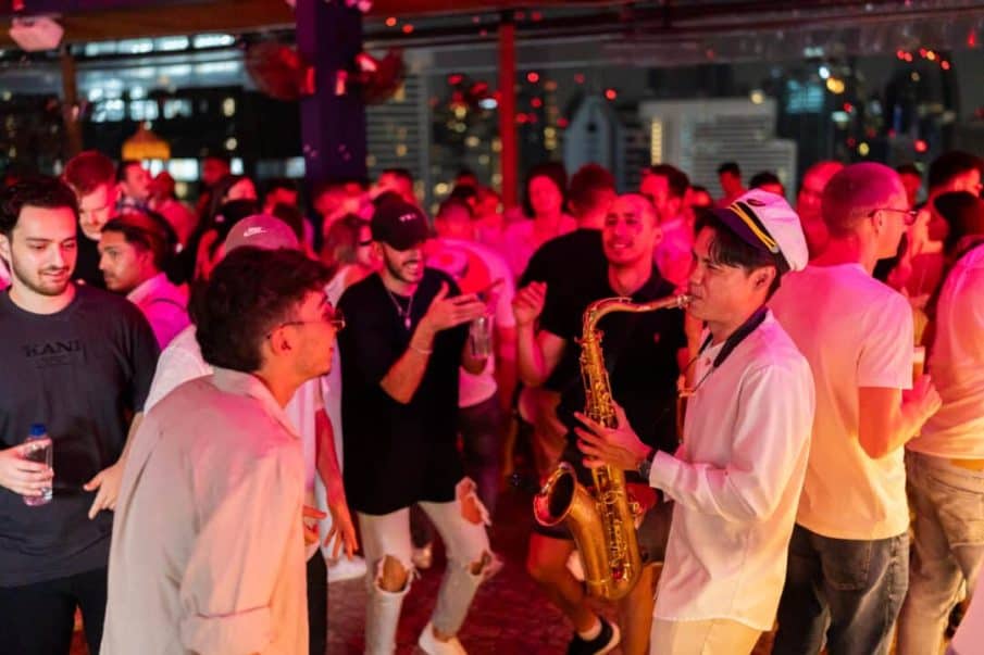 A birthday party photographer captures guests dancing around a saxophonist during a vibrant summer terrace club celebration.
