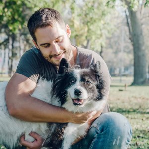 High-quality outdoor portrait of a smiling man sitting in a park and affectionately hugging his black and white border collie during a professional photo session.