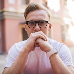 A professional urban portrait of a man with glasses and a white t-shirt, posing with his hands clasped in front of his face against a soft-focus city background.