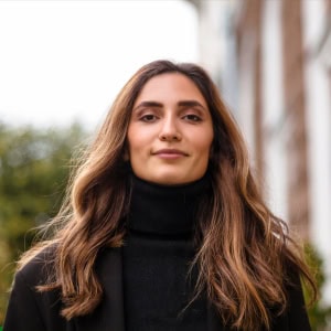 A professional outdoor portrait of a woman with long, wavy brown hair wearing a black turtleneck and coat, posing in front of a blurred architectural background in warsaw.