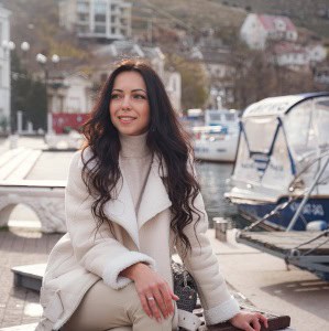 A woman with long dark hair wearing a white coat and beige turtleneck, smiling while sitting by a harbor with boats and buildings in the background.