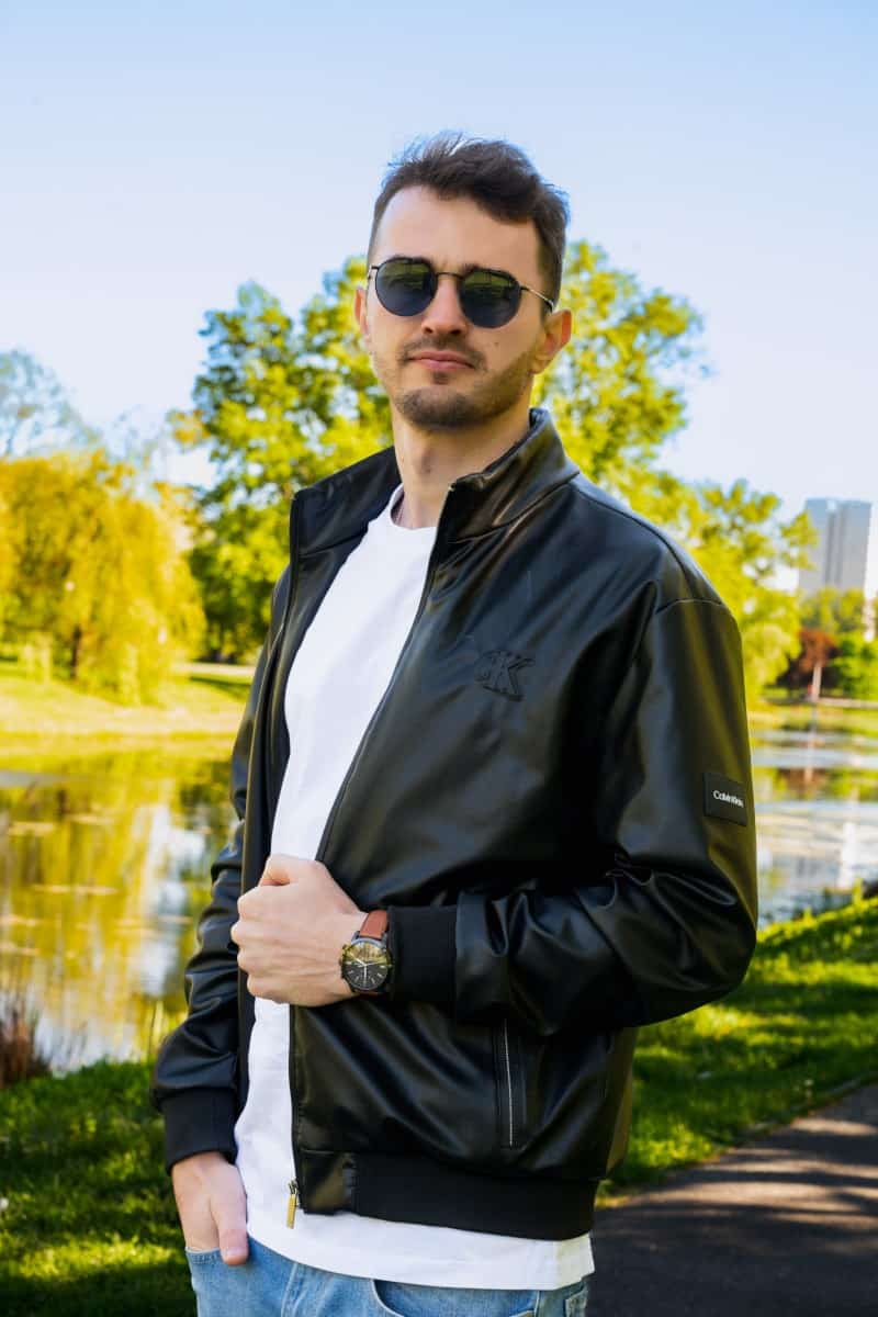 A man wearing a black calvin klein leather jacket and sunglasses posing in lazienki park in warsaw, with a lake and lush greenery in the background.