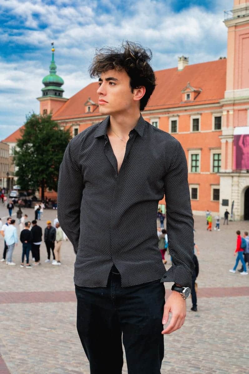 A young man in a black patterned shirt posing on castle square in warsaw's old town, with the royal castle and historic tenements in the background.