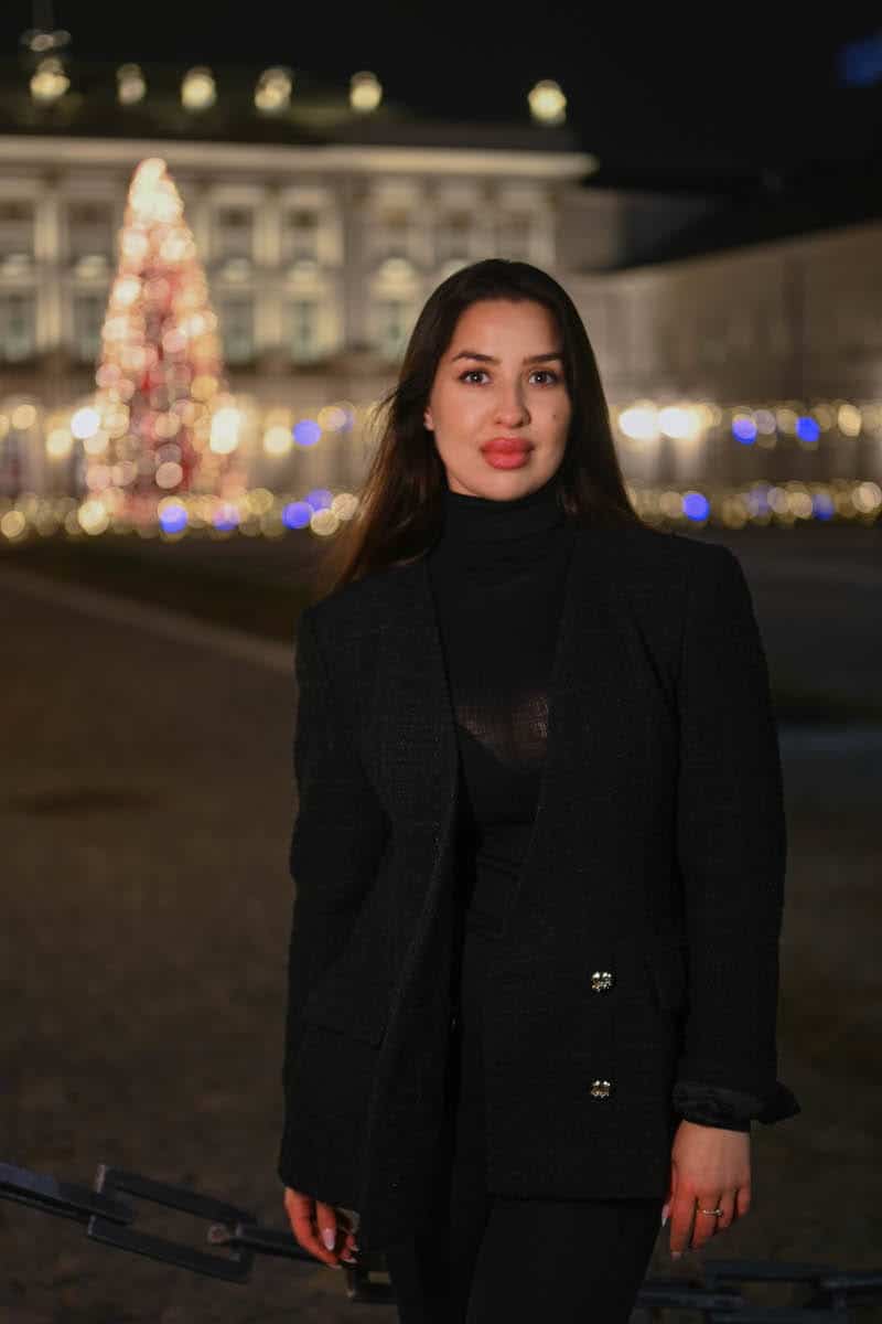 A woman in a black elegant blazer posing at night in front of the presidential palace in warsaw, with a glowing christmas tree and festive lights in the background.
