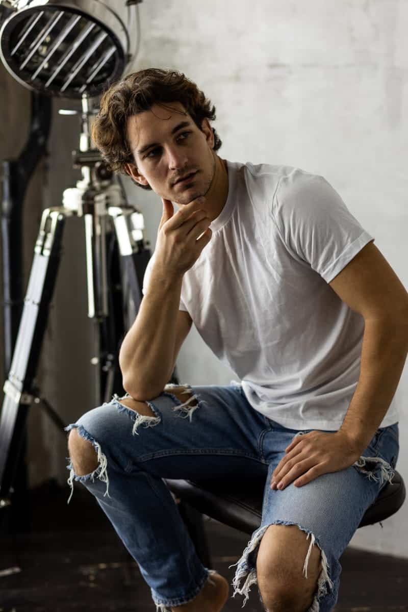 A man in a white t-shirt and distressed jeans sitting in a photo studio, posing thoughtfully against a professional studio light.