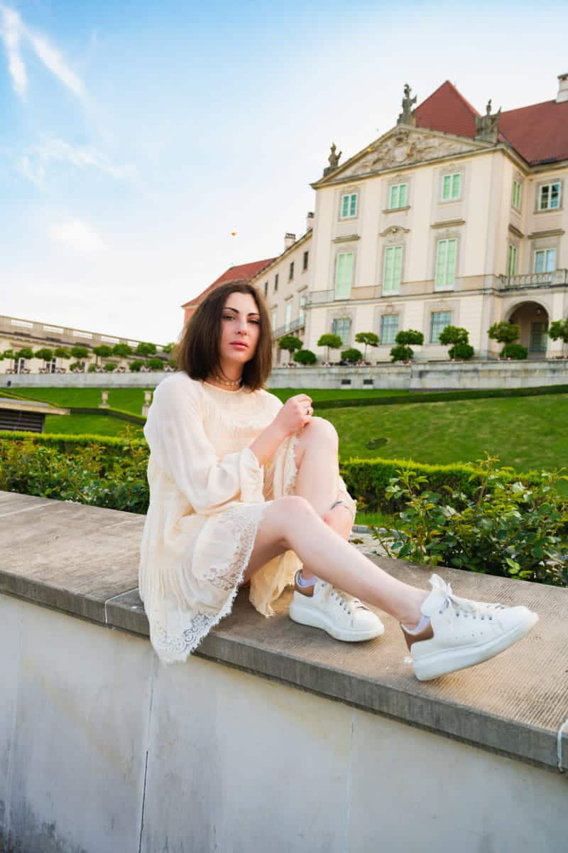 A woman in a white lace dress and sneakers sitting on a stone wall in the royal castle gardens in warsaw, with the historic castle building in the background.