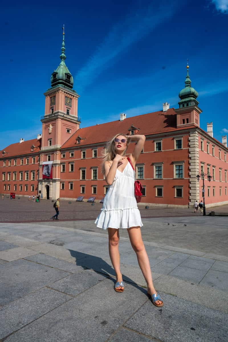 A woman in a white summer dress and sunglasses posing on castle square in front of the royal castle in warsaw on a sunny day.