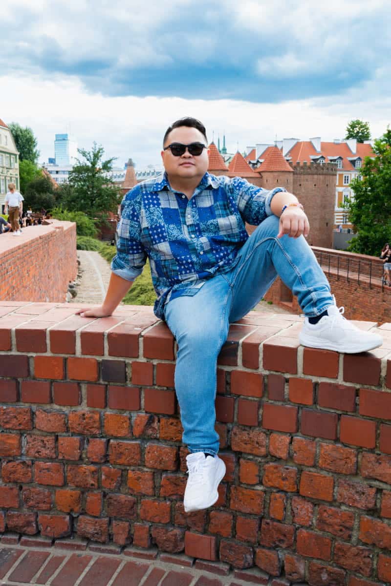 A man in a blue patterned shirt and jeans sitting on the historic red brick defensive walls of the warsaw barbican in the old town.