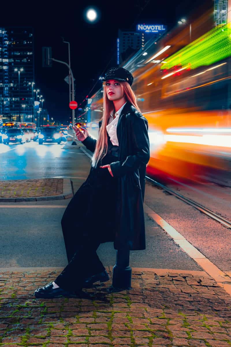 Professional urban night photography in warsaw city center, featuring a model leaning on a bollard near the novotel hotel with cinematic city lights and motion blur.