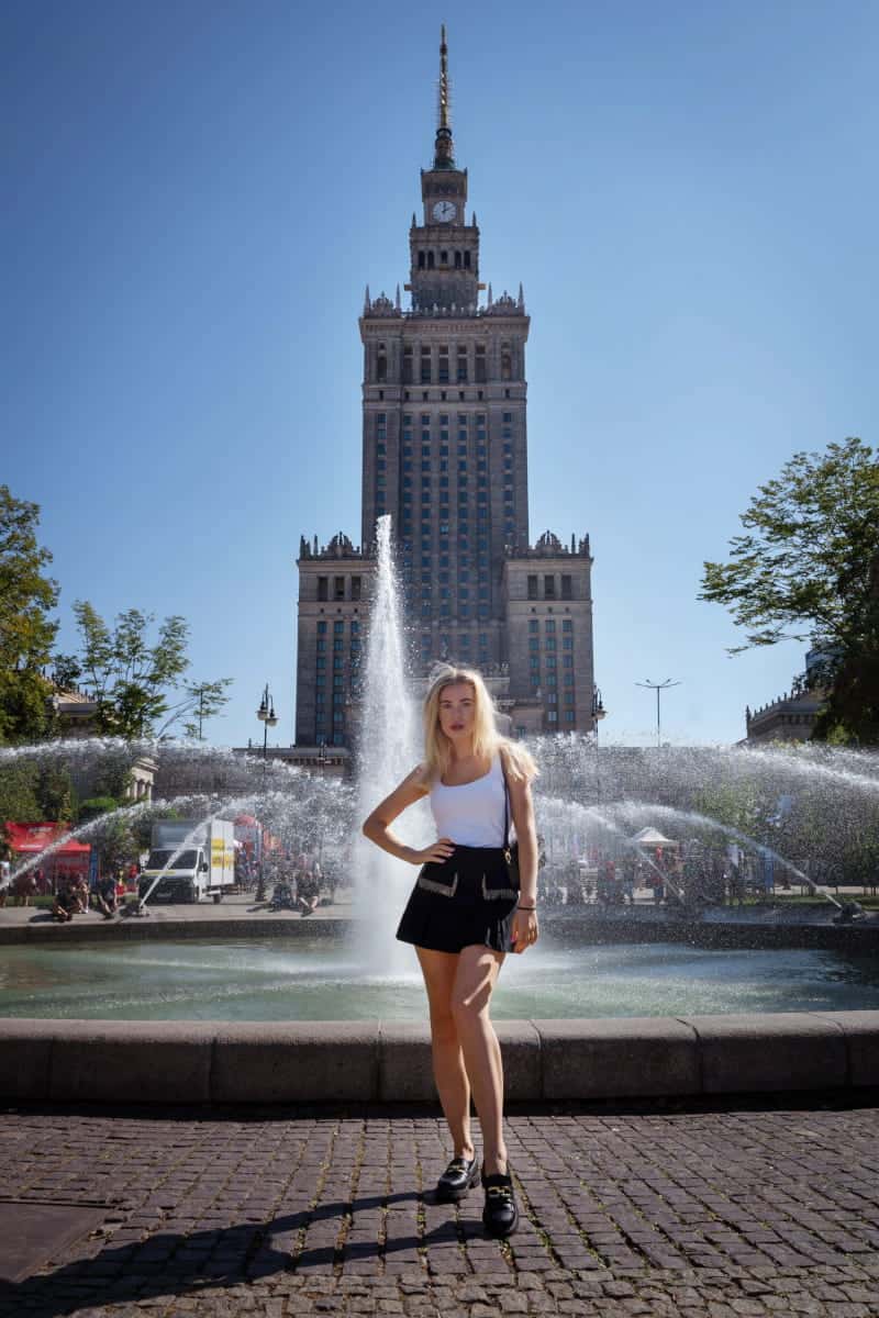Blonde woman posing in front of a fountain with the palace of culture and science in the background during an outdoor photo session in warsaw