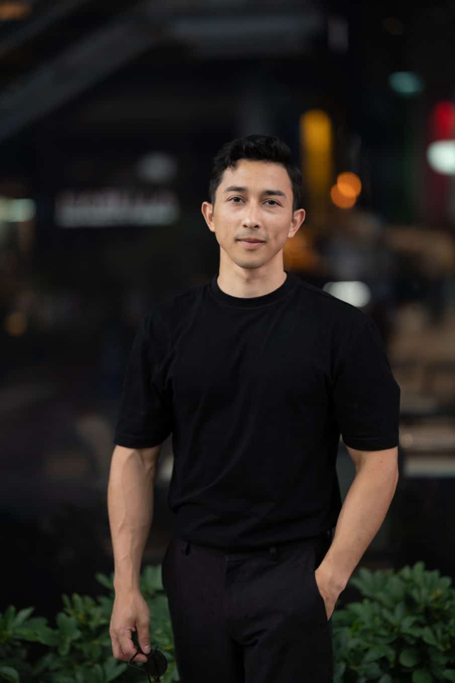 Portret męski lifestylowy z centrum warszawy. Mężczyzna w czarnym t-shircie i czarnych spodniach pozuje z ręką w kieszeni na tle rozmytych świateł miejskich. Minimalistyczna fotografia męska zrobiona wieczorem.