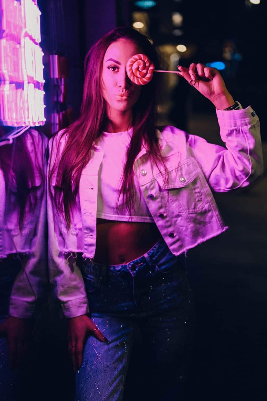 Portret kobiety w różowym, neonowym świetle, trzymającej lizaka. Kobieta ma na sobie białą kurtkę jeansową i pozuje w nocnym, miejskim plenerze. Zdjęcie lifestylowe z sesji beauty w kolorowym klimacie retro.