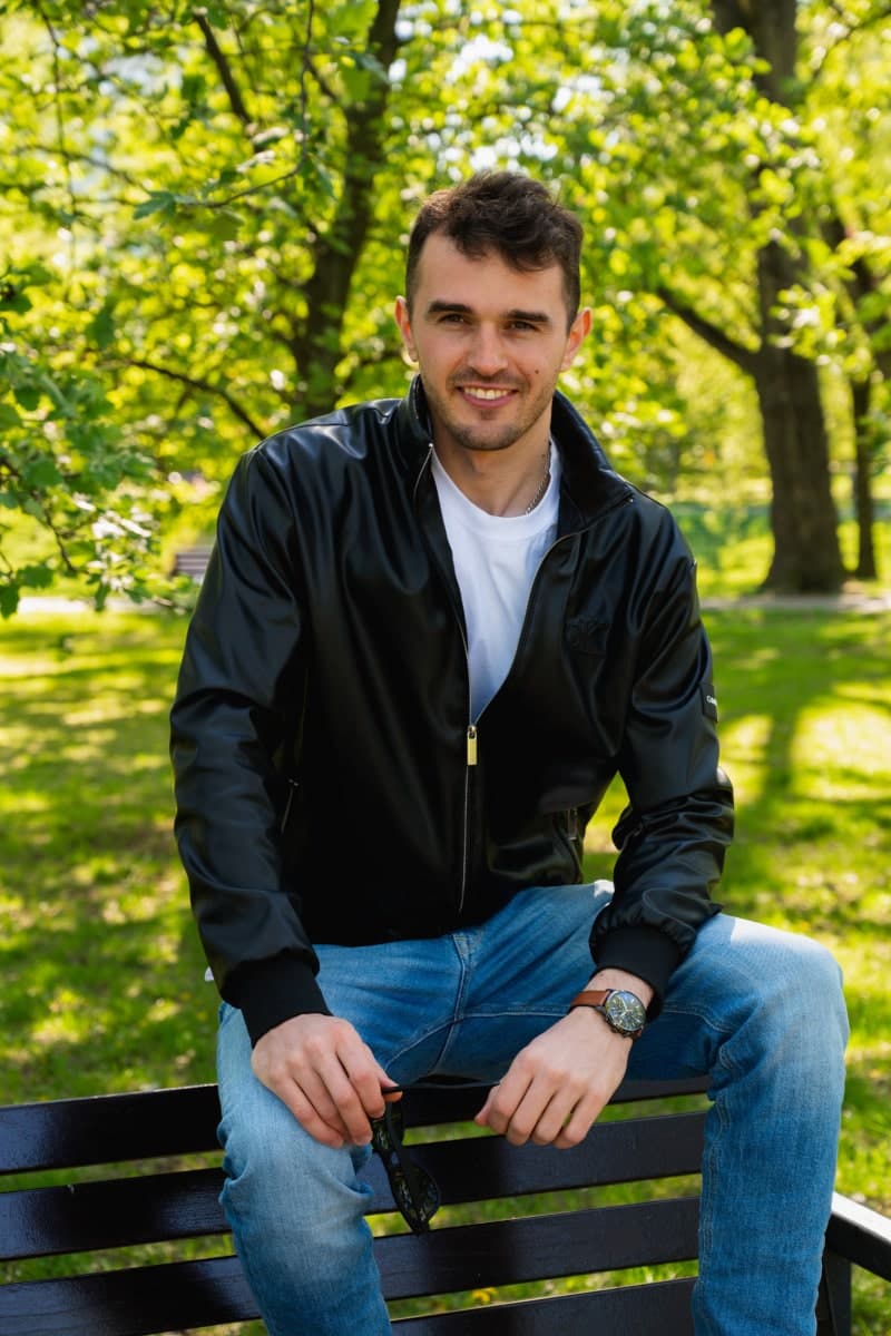 Uśmiechnięty portret męski wykonany w parku w warszawie, idealny do serwisu tinder. Mężczyzna ma na sobie czarną, błyszczącą kurtkę i biały t-shirt. Jasne, słoneczne zdjęcie w plenerze na tle intensywnie zielonych drzew.