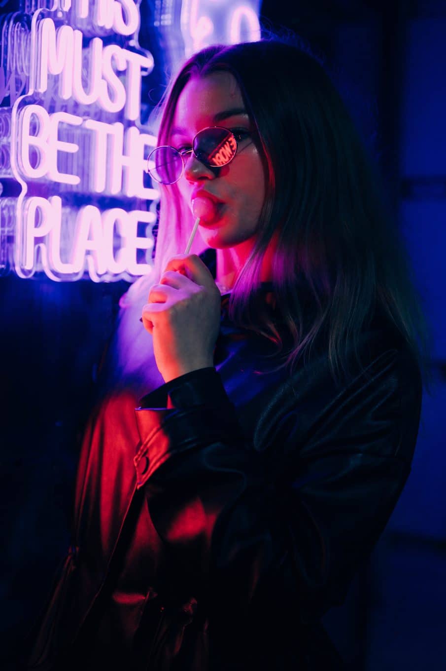 Dziewczyna w skórzanej kurtce i okularach, z lizakiem, na tle różowo-fioletowego neonu. Sesja tinder.