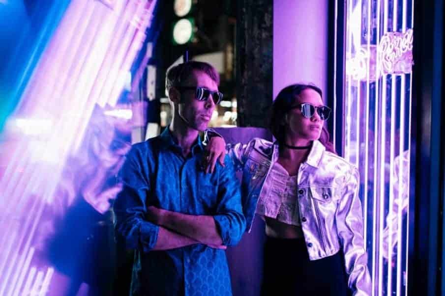 Portret pary w neonowym świetle, wykonany podczas miejskiej sesji zdjęciowej w warszawie. Mężczyzna w niebieskiej koszuli i kobieta w srebrnej kurtce pozują w okularach przeciwsłonecznych na tle fioletowych neonów. Romantyczna sesja zdjęciowa w nocnym, miejskim klimacie.