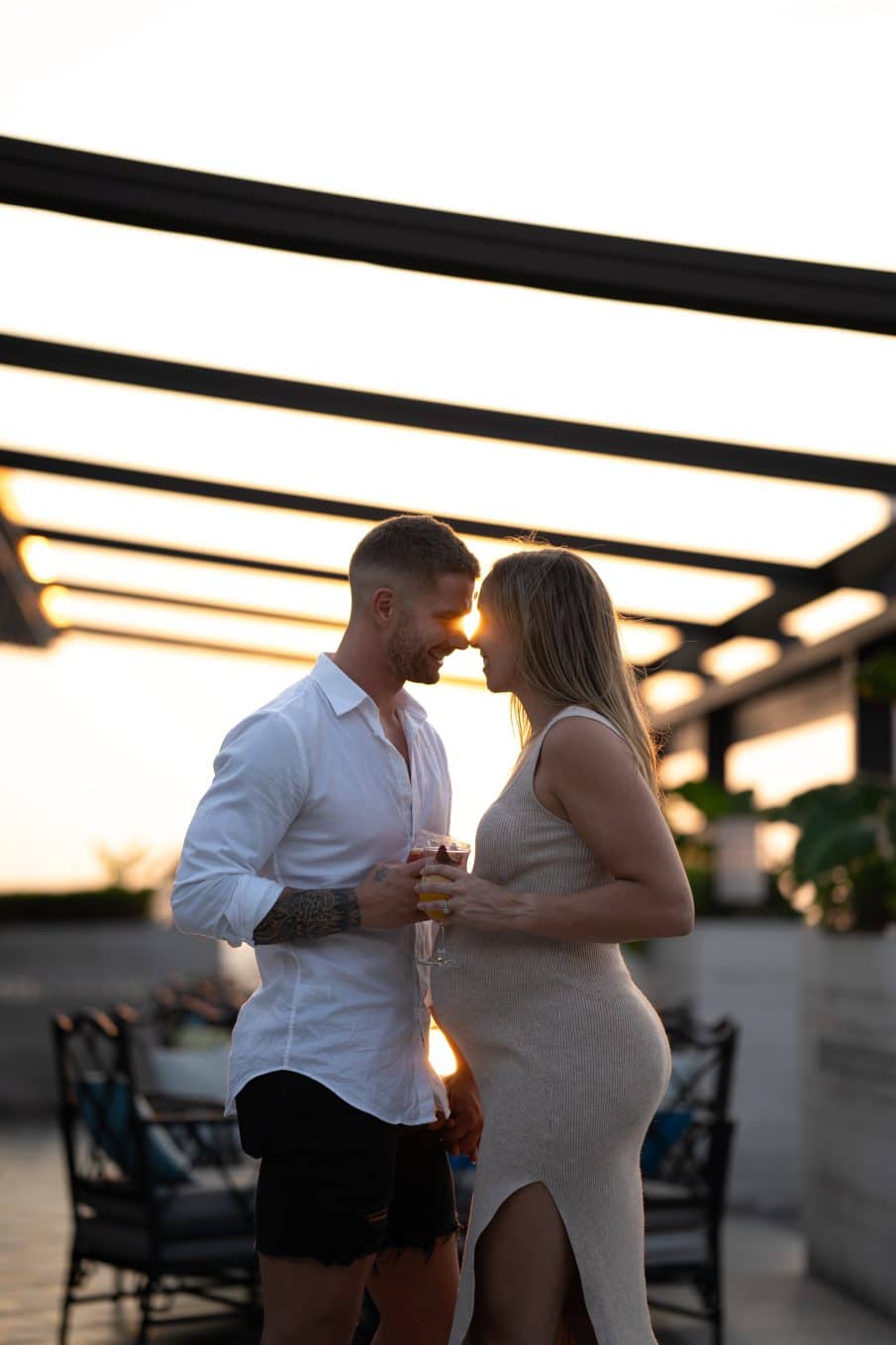 Romantyczna sesja zdjęciowa pary w świetle zachodzącego słońca na tarasie widokowym w warszawie. Mężczyzna w białej koszuli i kobieta w beżowej sukience (w ciąży), trzymający drinki. Intymny portret pary w blasku złotego słońca.