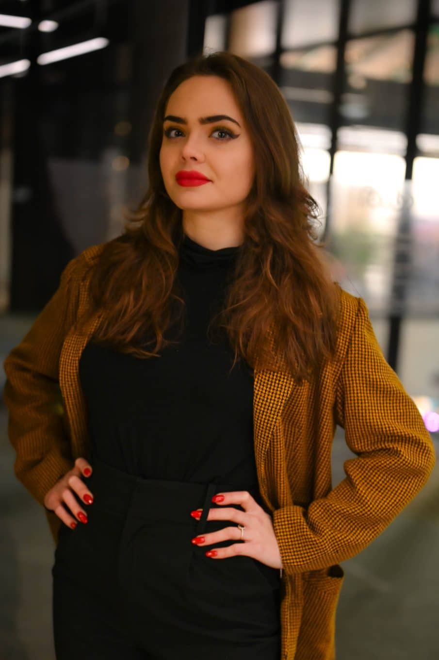 Profesjonalny portret biznesowy kobiety z długimi, brązowymi włosami, stojącej z rękami na biodrach. Ma na sobie czarny golf, czarne spodnie i musztardową marynarkę w kratkę. Mocny akcent to czerwona szminka i manicure. Zdjęcie wykonane w warszawie.