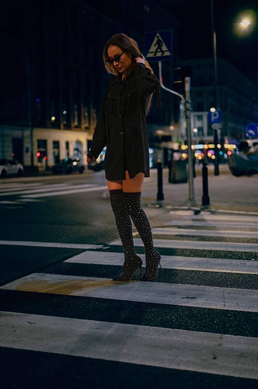Nocny portret kobiecy w miejskim stylu. Kobieta w czarnej koszuli z frędzlami i okularach przeciwsłonecznych pozuje na ulicy warszawy, na tle znaku drogowego i rozmytych świateł miasta. Zdjęcie lifestylowe z nocnej sesji w centrum.