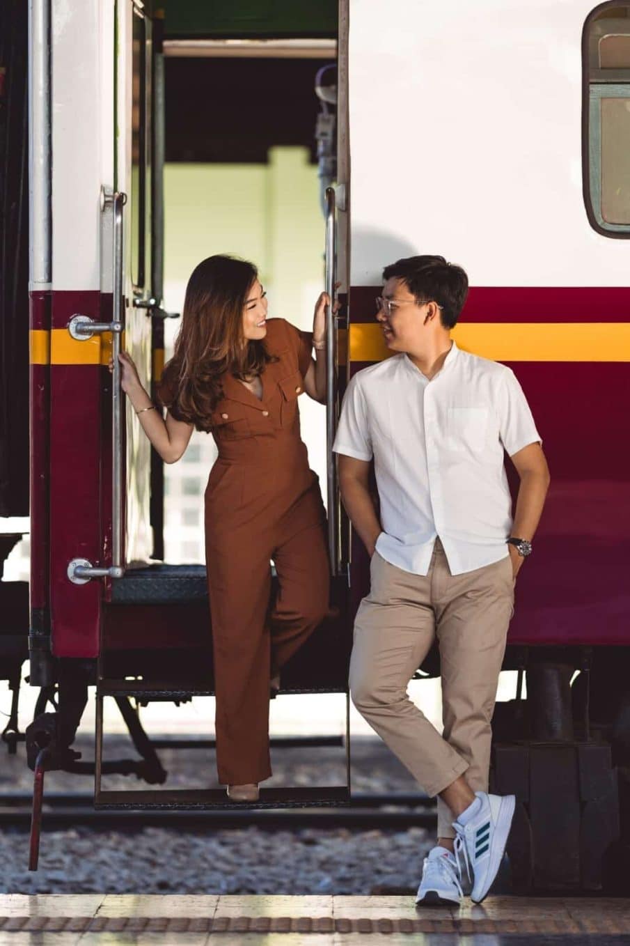 Sesja dla par wykonana na dworcu kolejowym, przy wejściu do pociągu. Kobieta w brązowym kombinezonie i mężczyzna w białej koszuli patrzą na siebie z uśmiechem. Romantyczna sesja zdjęciowa w miejskim, industrialnym klimacie.