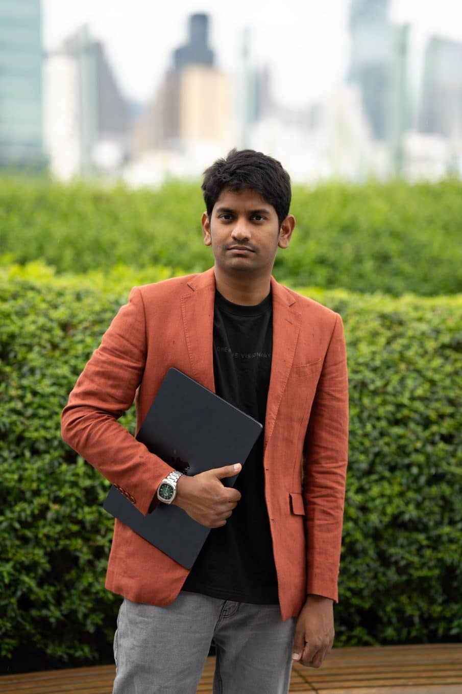 Biznesowe zdjęcie na linkedin mężczyzny w ceglanej marynarce i czarnej koszulce, trzymającego laptopa. Ujęcie portretowe z poważnym wyrazem twarzy na tle zielonych krzewów.